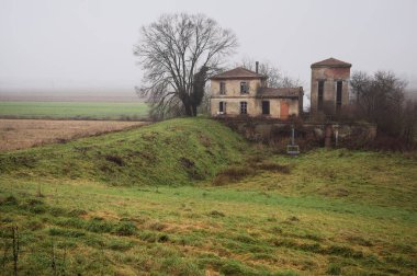 Bir tarlada terk edilmiş bir baraj, yanında çıplak bir ağaç olan bir hendek, İtalya 'nın kışın kırsalında sisli ve bulutlu bir günde.