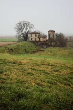 Bir tarlada terk edilmiş bir baraj, yanında çıplak bir ağaç olan bir hendek, İtalya 'nın kışın kırsalında sisli ve bulutlu bir günde.