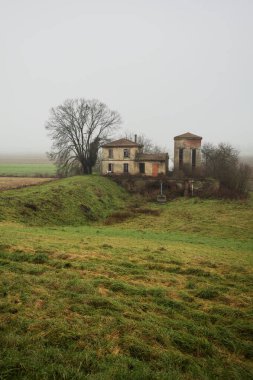 Bir tarlada terk edilmiş bir baraj, yanında çıplak bir ağaç olan bir hendek, İtalya 'nın kışın kırsalında sisli ve bulutlu bir günde.