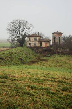 Bir tarlada terk edilmiş bir baraj, yanında çıplak bir ağaç olan bir hendek, İtalya 'nın kışın kırsalında sisli ve bulutlu bir günde.