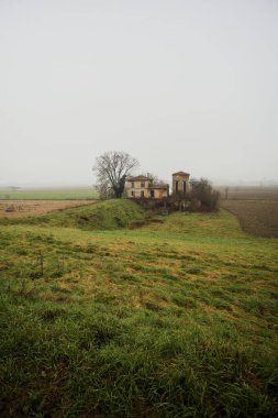 Bir tarlada terk edilmiş bir baraj, yanında çıplak bir ağaç olan bir hendek, İtalya 'nın kışın kırsalında sisli ve bulutlu bir günde.