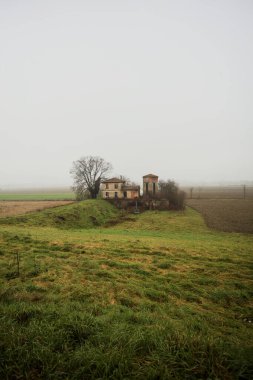 Bir tarlada terk edilmiş bir baraj, yanında çıplak bir ağaç olan bir hendek, İtalya 'nın kışın kırsalında sisli ve bulutlu bir günde.