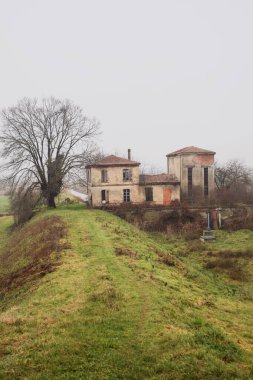 Abandoned dam building in a field, framing a ditch with water, with a bare tree next to it, on a misty and cloudy day in the italian countryside in winter