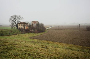 Bir tarlada terk edilmiş bir baraj, yanında çıplak bir ağaç olan bir hendek, İtalya 'nın kışın kırsalında sisli ve bulutlu bir günde.
