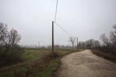 Çamurlu bir zeminde toprak izi, tepesinde bir viraj oluşturur, kıvrımın ucunda tuğla ve beton bir kulübe ile çevrili ağaçlar, ve diğer ucunda tahta direklerde elektrik hattı, İtalya kırsalında bulutlu ve sisli bir günde, kış mevsiminde
