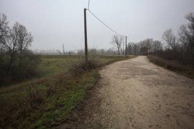 Çamurlu bir zeminde toprak izi, tepesinde bir viraj oluşturur, kıvrımın ucunda tuğla ve beton bir kulübe ile çevrili ağaçlar, ve diğer ucunda tahta direklerde elektrik hattı, İtalya kırsalında bulutlu ve sisli bir günde, kış mevsiminde