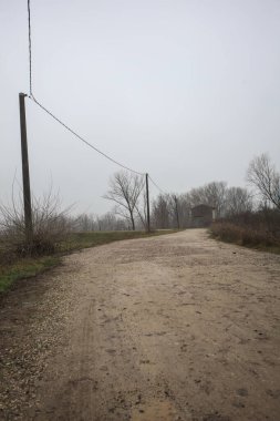 Çamurlu bir zeminde toprak izi, tepesinde bir viraj oluşturur, kıvrımın ucunda tuğla ve beton bir kulübe ile çevrili ağaçlar, ve diğer ucunda tahta direklerde elektrik hattı, İtalya kırsalında bulutlu ve sisli bir günde, kış mevsiminde