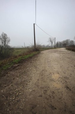 Çamurlu bir zeminde toprak izi, tepesinde bir viraj oluşturur, kıvrımın ucunda tuğla ve beton bir kulübe ile çevrili ağaçlar, ve diğer ucunda tahta direklerde elektrik hattı, İtalya kırsalında bulutlu ve sisli bir günde, kış mevsiminde
