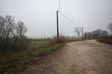 Çamurlu bir zeminde toprak izi, tepesinde bir viraj oluşturur, kıvrımın ucunda tuğla ve beton bir kulübe ile çevrili ağaçlar, ve diğer ucunda tahta direklerde elektrik hattı, İtalya kırsalında bulutlu ve sisli bir günde, kış mevsiminde