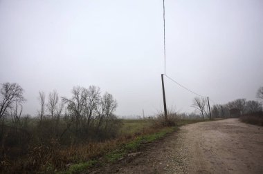 Çamurlu bir zeminde toprak izi, tepesinde bir viraj oluşturur, kıvrımın ucunda tuğla ve beton bir kulübe ile çevrili ağaçlar, ve diğer ucunda tahta direklerde elektrik hattı, İtalya kırsalında bulutlu ve sisli bir günde, kış mevsiminde