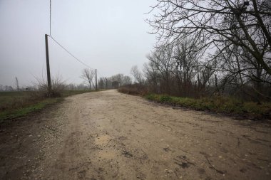 Çamurlu bir zeminde toprak izi, tepesinde bir viraj oluşturur, kıvrımın ucunda tuğla ve beton bir kulübe ile çevrili ağaçlar, ve diğer ucunda tahta direklerde elektrik hattı, İtalya kırsalında bulutlu ve sisli bir günde, kış mevsiminde