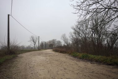 Çamurlu bir zeminde toprak izi, tepesinde bir viraj oluşturur, kıvrımın ucunda tuğla ve beton bir kulübe ile çevrili ağaçlar, ve diğer ucunda tahta direklerde elektrik hattı, İtalya kırsalında bulutlu ve sisli bir günde, kış mevsiminde