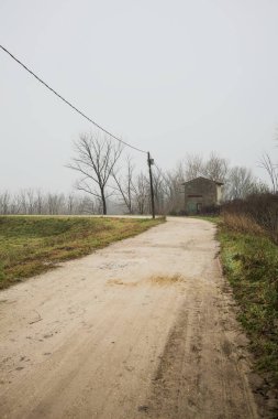 Çamurlu bir zeminde toprak izi, tepesinde bir viraj oluşturur, kıvrımın ucunda tuğla ve beton bir kulübe ile çevrili ağaçlar, ve diğer ucunda tahta direklerde elektrik hattı, İtalya kırsalında bulutlu ve sisli bir günde, kış mevsiminde