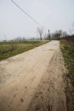 Çamurlu bir zeminde toprak izi, tepesinde bir viraj oluşturur, kıvrımın ucunda tuğla ve beton bir kulübe ile çevrili ağaçlar, ve diğer ucunda tahta direklerde elektrik hattı, İtalya kırsalında bulutlu ve sisli bir günde, kış mevsiminde