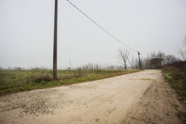 Çamurlu bir zeminde toprak izi, tepesinde bir viraj oluşturur, kıvrımın ucunda tuğla ve beton bir kulübe ile çevrili ağaçlar, ve diğer ucunda tahta direklerde elektrik hattı, İtalya kırsalında bulutlu ve sisli bir günde, kış mevsiminde