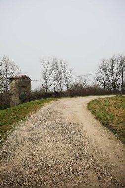 Çamurlu bir zeminde toprak izi, tepesinde bir viraj oluşturur, kıvrımın ucunda tuğla ve beton bir kulübe ile çevrili ağaçlar, ve diğer ucunda tahta direklerde elektrik hattı, İtalya kırsalında bulutlu ve sisli bir günde, kış mevsiminde