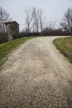 Çamurlu bir zeminde toprak izi, tepesinde bir viraj oluşturur, kıvrımın ucunda tuğla ve beton bir kulübe ile çevrili ağaçlar, ve diğer ucunda tahta direklerde elektrik hattı, İtalya kırsalında bulutlu ve sisli bir günde, kış mevsiminde