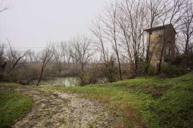 Bataklıkta bir havzanın kenarındaki toprak yol, kıyısında ağaçlar olan, ve havzayı çevreleyen beton bir barakanın tepesinde, İtalya 'nın kışın kırsalında bulutlu ve sisli bir günde