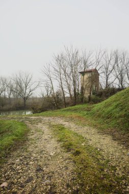 Bataklıkta bir havzanın kenarındaki toprak yol, kıyısında ağaçlar olan, ve havzayı çevreleyen beton bir barakanın tepesinde, İtalya 'nın kışın kırsalında bulutlu ve sisli bir günde