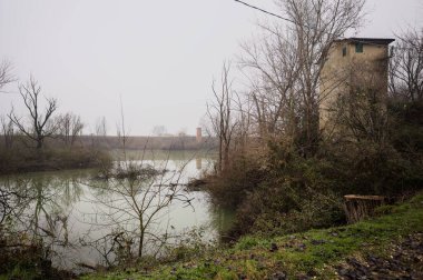 Bir bataklıkta, çıplak ağaçlarla çevrili bir havzada ve karşı tarafında bir baraj var, tepesinde de bir kulübe, hepsi İtalya 'nın kışın kırsalında bulutlu ve sisli bir günde suya yansıyor.