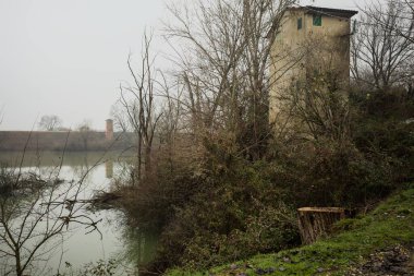 Bir bataklıkta, çıplak ağaçlarla çevrili bir havzada ve karşı tarafında bir baraj var, tepesinde de bir kulübe, hepsi İtalya 'nın kışın kırsalında bulutlu ve sisli bir günde suya yansıyor.