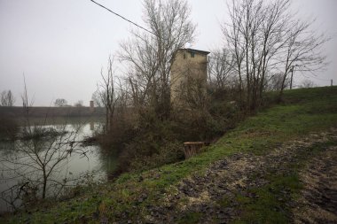 Bataklıkta bir havzanın kenarındaki toprak yol, kıyısında ağaçlar olan, ve havzayı çevreleyen beton bir barakanın tepesinde, İtalya 'nın kışın kırsalında bulutlu ve sisli bir günde