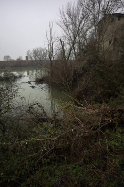 Bir bataklıkta, çıplak ağaçlarla çevrili bir havzada ve karşı tarafında bir baraj var, tepesinde de bir kulübe, hepsi İtalya 'nın kışın kırsalında bulutlu ve sisli bir günde suya yansıyor.