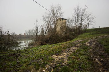 Bataklıkta bir havzanın kenarındaki toprak yol, kıyısında ağaçlar olan, ve havzayı çevreleyen beton bir barakanın tepesinde, İtalya 'nın kışın kırsalında bulutlu ve sisli bir günde