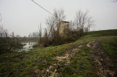 Bataklıkta bir havzanın kenarındaki toprak yol, kıyısında ağaçlar olan, ve havzayı çevreleyen beton bir barakanın tepesinde, İtalya 'nın kışın kırsalında bulutlu ve sisli bir günde
