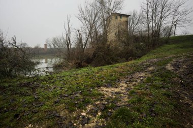 Bataklıkta bir havzanın kenarındaki toprak yol, kıyısında ağaçlar olan, ve havzayı çevreleyen beton bir barakanın tepesinde, İtalya 'nın kışın kırsalında bulutlu ve sisli bir günde