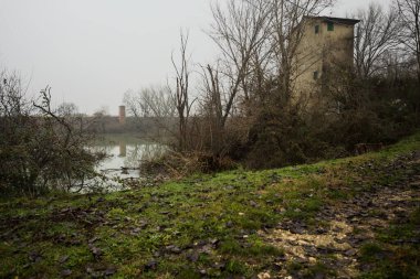 Bataklıkta bir havzanın kenarındaki toprak yol, kıyısında ağaçlar olan, ve havzayı çevreleyen beton bir barakanın tepesinde, İtalya 'nın kışın kırsalında bulutlu ve sisli bir günde