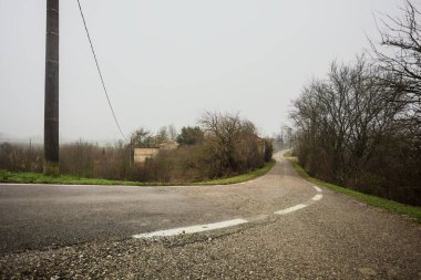 Kışın İtalya kırsalında bulutlu ve sisli bir günde, yol kenarında bir malikanesi olan küçük bir koruluktan geçerek setin tepesine inen bir yol.
