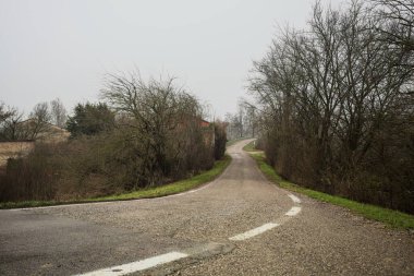 Kışın İtalya kırsalında bulutlu ve sisli bir günde, yol kenarında bir malikanesi olan küçük bir koruluktan geçerek setin tepesine inen bir yol.