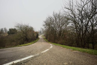 Kışın İtalya kırsalında bulutlu ve sisli bir günde, yol kenarında bir malikanesi olan küçük bir koruluktan geçerek setin tepesine inen bir yol.