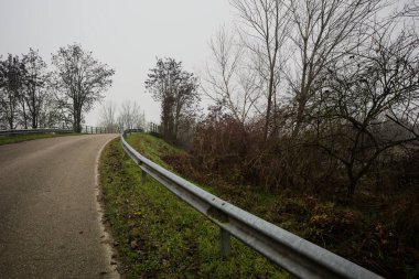 Kışın İtalya kırsalında sisli bir günde, bir su deresi, çıplak ağaçlar ve uzun otlarla çevrili bir viraj yaparak setin üzerindeki yola tırmanmak.