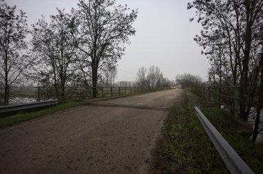 Sisli ve bulutlu bir günde, İtalya kırsalında köprünün diğer ucunda, arka planda bir malikane bulunan çıplak ağaçlar tarafından çerçevelenmiş kısmen yıpranmış yol köprüsü.