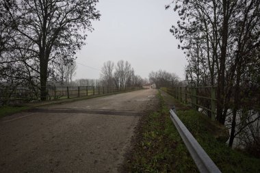 Sisli ve bulutlu bir günde, İtalya kırsalında köprünün diğer ucunda, arka planda bir malikane bulunan çıplak ağaçlar tarafından çerçevelenmiş kısmen yıpranmış yol köprüsü.