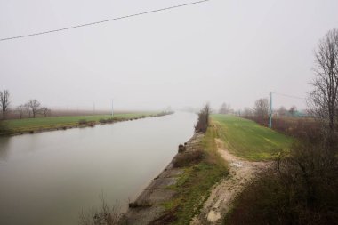 İtalya 'nın kırsal kesimlerinde sisli ve bulutlu bir günde sulara yansıyan ağaçlarla çevrili tarlalar arasındaki nehir.