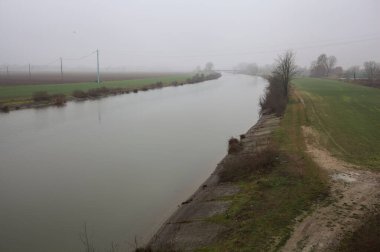 İtalya 'nın kırsal kesimlerinde sisli ve bulutlu bir günde sulara yansıyan ağaçlarla çevrili tarlalar arasındaki nehir.