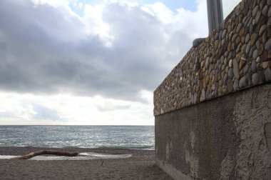 Bulutlu ve rüzgarlı bir günde, kayalar ve taş bir duvarla çerçevelenmiş boş bir plaj.