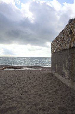 Bulutlu ve rüzgarlı bir günde, kayalar ve taş bir duvarla çerçevelenmiş boş bir plaj.