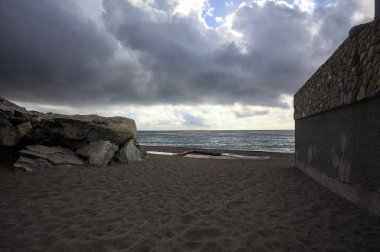 Bulutlu ve rüzgarlı bir günde, kayalar ve taş bir duvarla çerçevelenmiş boş bir plaj.
