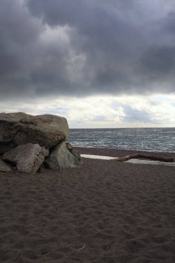 Bulutlu ve rüzgarlı bir günde, kayalar ve taş bir duvarla çerçevelenmiş boş bir plaj.