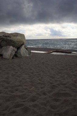 Bulutlu ve rüzgarlı bir günde, kayalar ve taş bir duvarla çerçevelenmiş boş bir plaj.