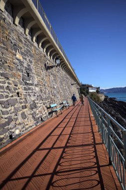 Taştan bir duvarla çevrili bir gezinti güvertesi ve güneşli bir günde gölgesini kayalıkların üzerine düşüren bir korkuluk.