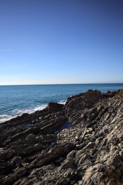 Denizin dalgaları bir uçurumun kayalıklarına çarpıyor. Güneş ışığı yukarıdan görünen suya vuruyor.