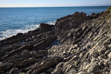 Denizin dalgaları bir uçurumun kayalıklarına çarpıyor. Güneş ışığı yukarıdan görünen suya vuruyor.