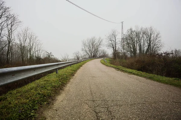 Kışın İtalya kırsalında sisli bir günde, bir su deresi, çıplak ağaçlar ve uzun otlarla çevrili bir viraj yaparak setin üzerindeki yola tırmanmak.