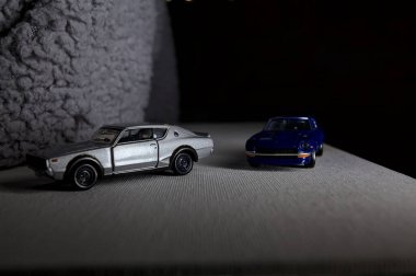 Mantova, İtalya - 3RD JANUARY 2025 Takara Tomy kopyaları gri Nissan Skyline 2000 GT-R ve mavi Nissan Fairlady Z beyaz bir panoda, gri bir kumaş parçasının yanında, uçurumun kenarındaki bir dağ yolunda yarış yapıyormuş gibi yapıyor.