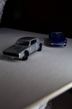 Mantova, İtalya - 3RD JANUARY 2025 Takara Tomy kopyaları gri Nissan Skyline 2000 GT-R ve mavi Nissan Fairlady Z beyaz bir panoda, gri bir kumaş parçasının yanında, uçurumun kenarındaki bir dağ yolunda yarış yapıyormuş gibi yapıyor.